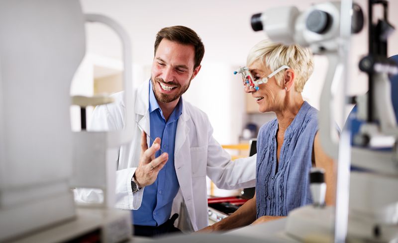 OphthoLegal A friendly male optometrist explaining vision correction options to a senior female patient wearing a trial frame.